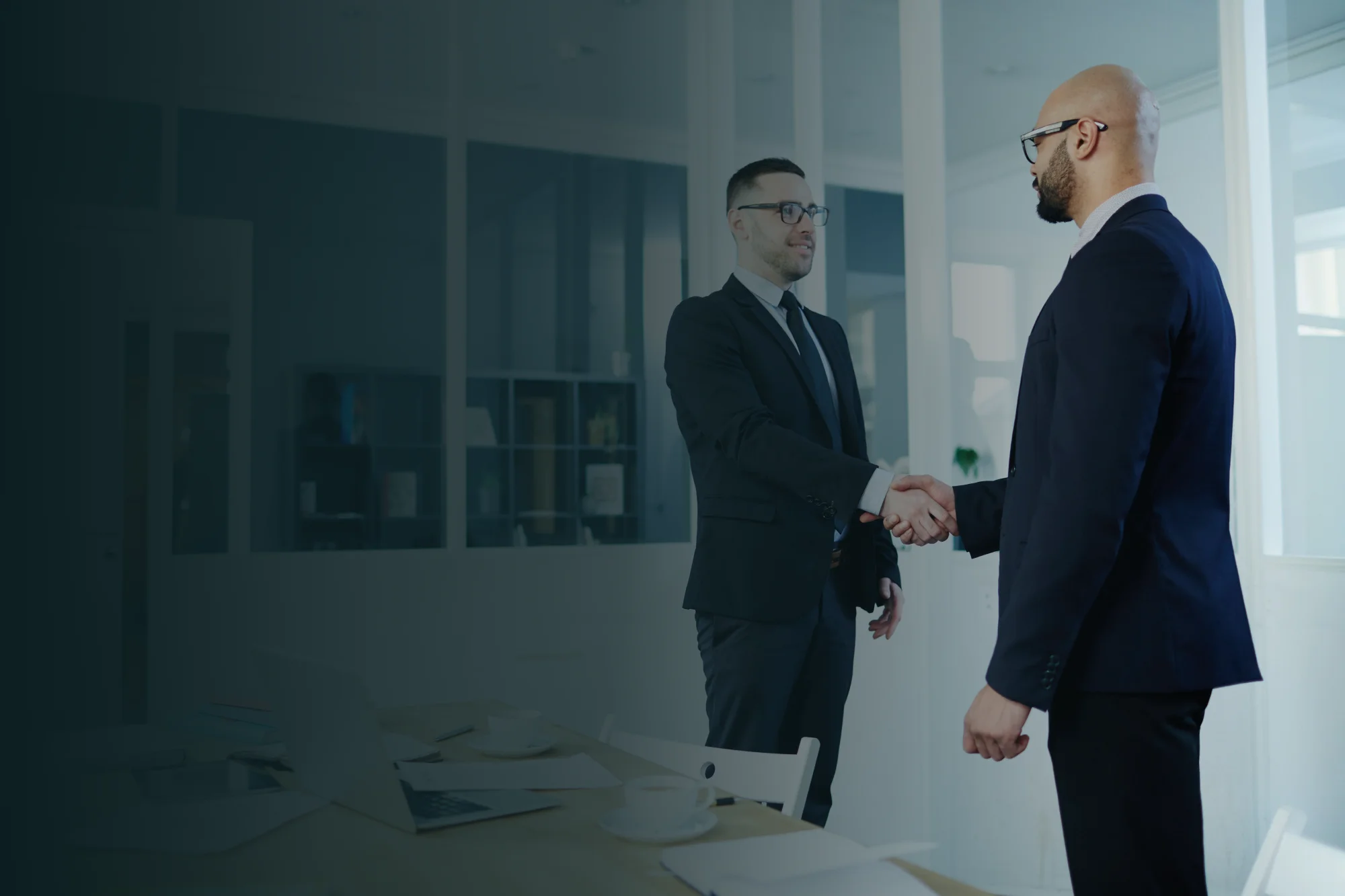 Two professional men in suits shaking hands in a modern, brightly lit office space. The left side of the image features a dark blue gradient overlay, suggesting a professional partnership or a successful business agreement.